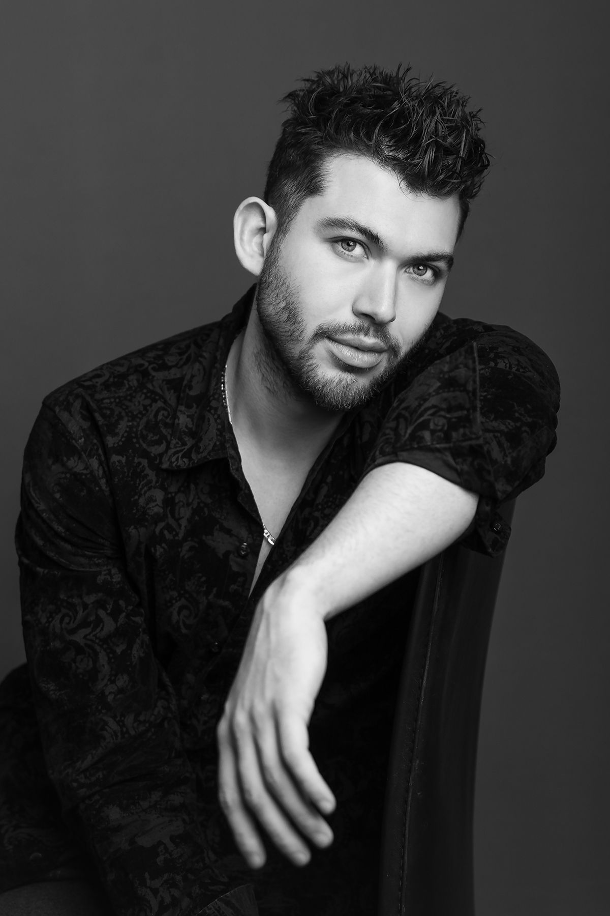 Black and white studio portrait of a bearded man in dark patterned shirt leaning on chair armrest.
