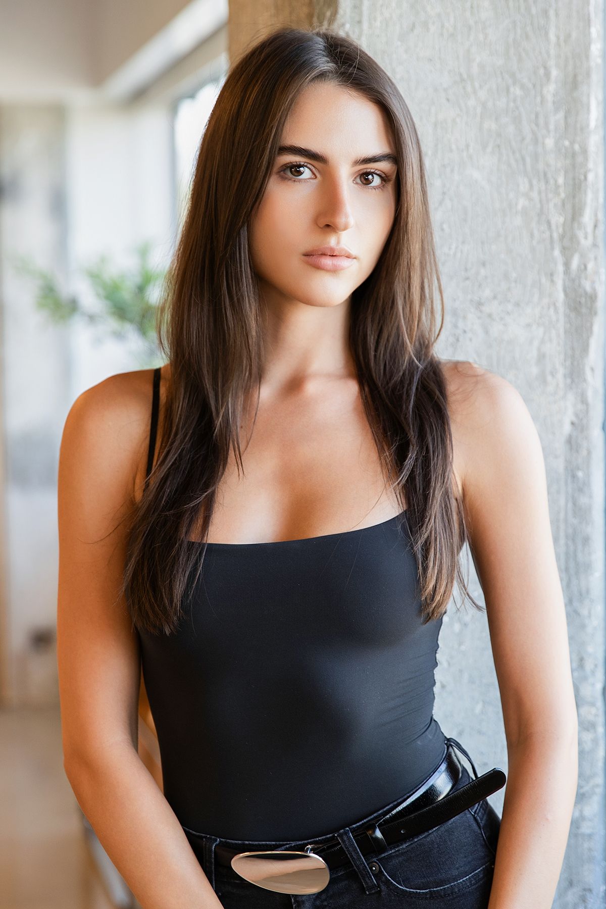 Young woman with long dark hair wearing a black spaghetti strap bodysuit and jeans, leaning against a concrete pillar.