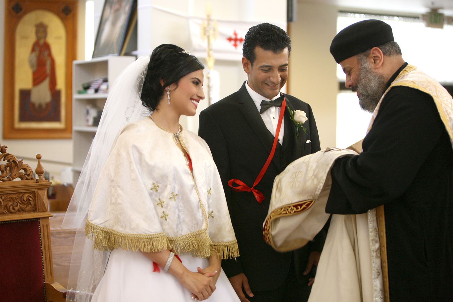 Smiling bride in white gown and veil stands with groom during a traditional Coptic Orthodox church wedding.