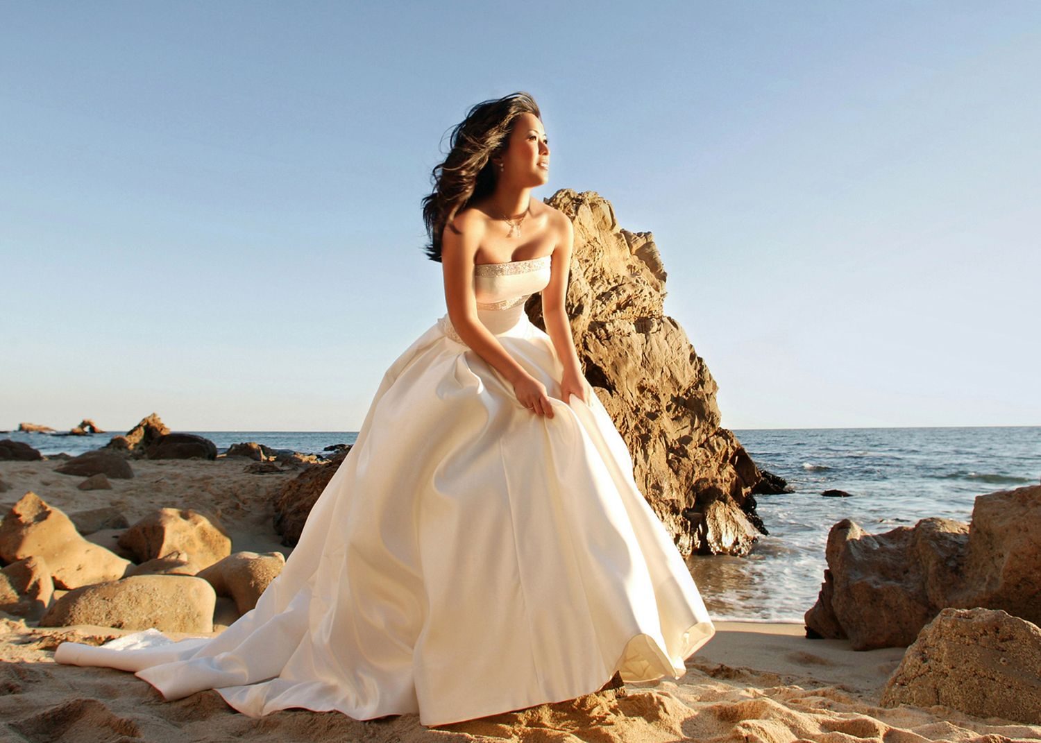 Bride in elegant white ball gown posing on rocky beach with ocean backdrop on a clear sunny day.
