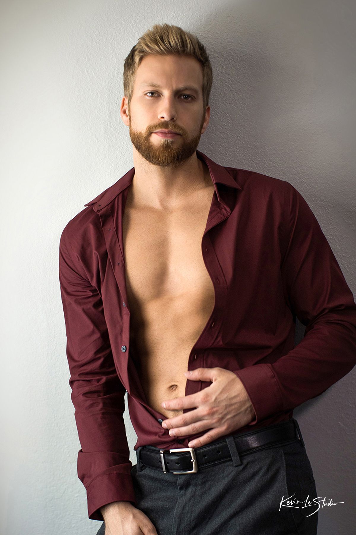 Muscular man with beard wearing open burgundy shirt and black pants posing confidently against white wall.