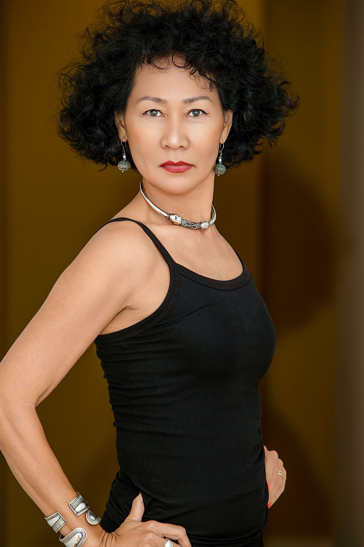 A confident woman in a black tank top with curly hair, silver jewelry, and red lips poses with hands on hips.