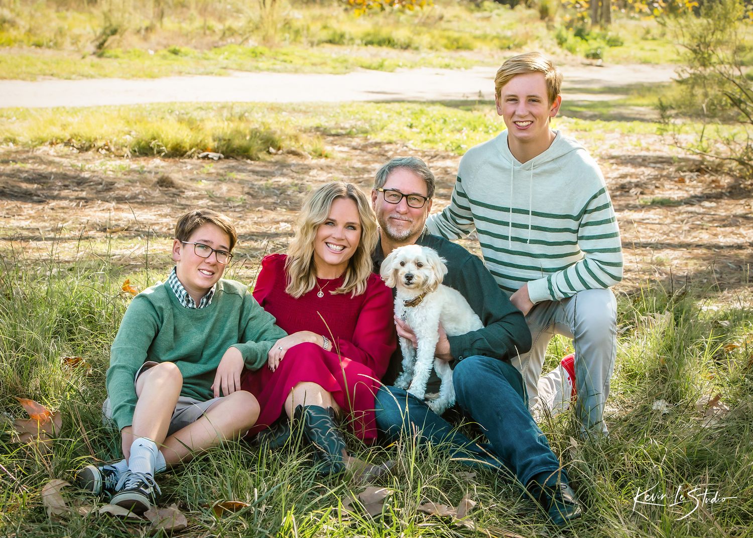 Happy family of four with a white fluffy dog sitting together outdoors in a sunny park setting.