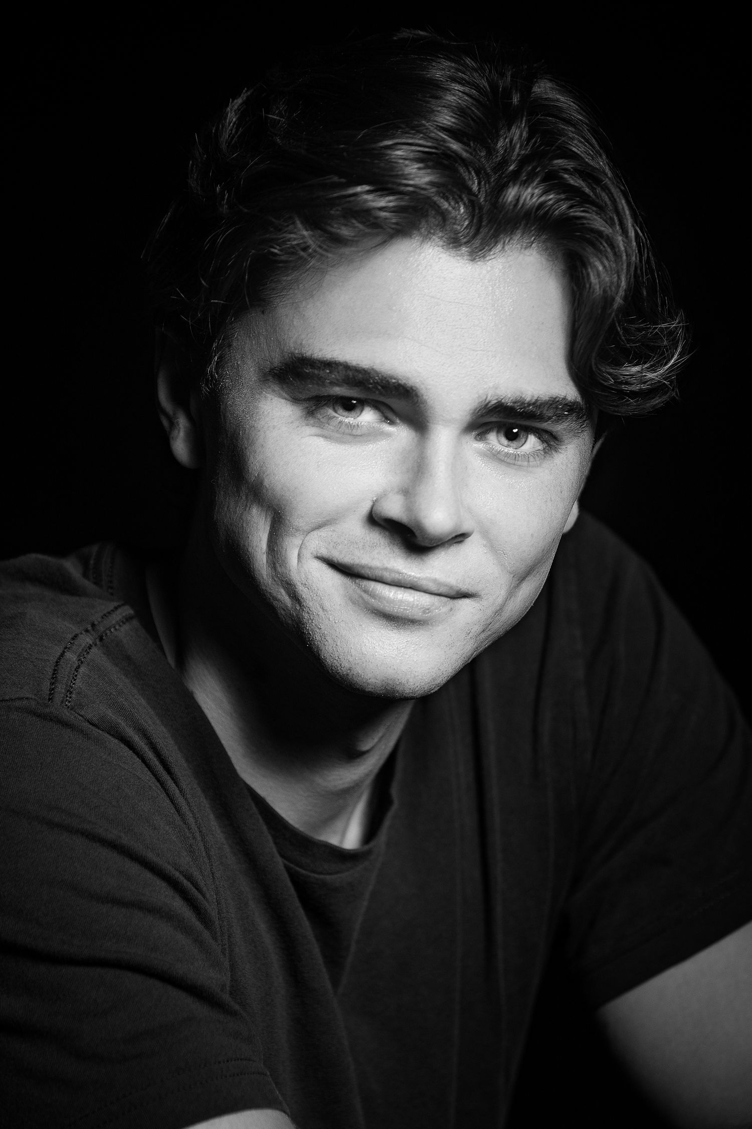 Black and white portrait of a young smiling man with wavy hair wearing a dark t-shirt against black background.