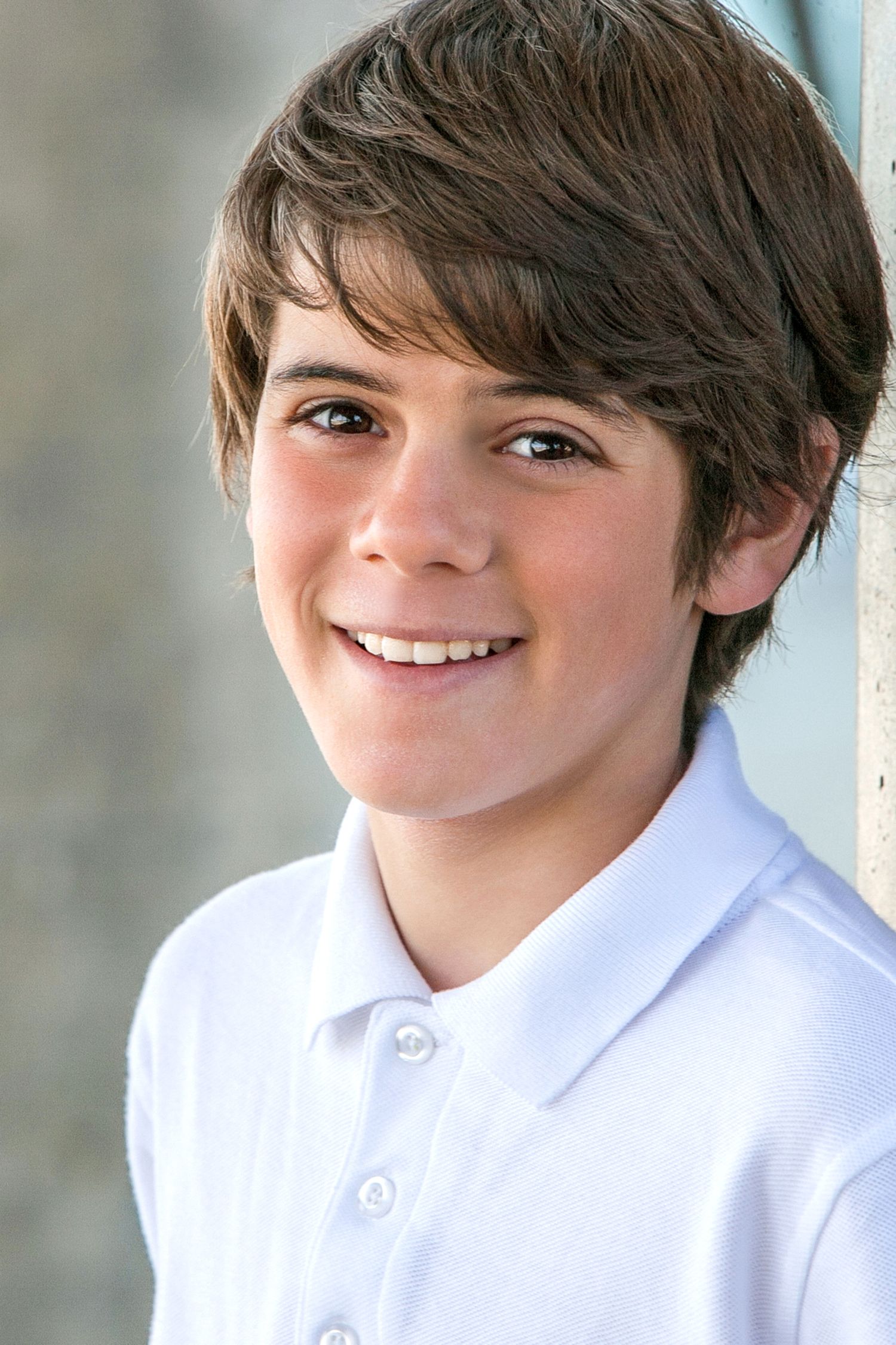 Smiling young boy with brown hair wearing a white polo shirt poses for a portrait photo outdoors.