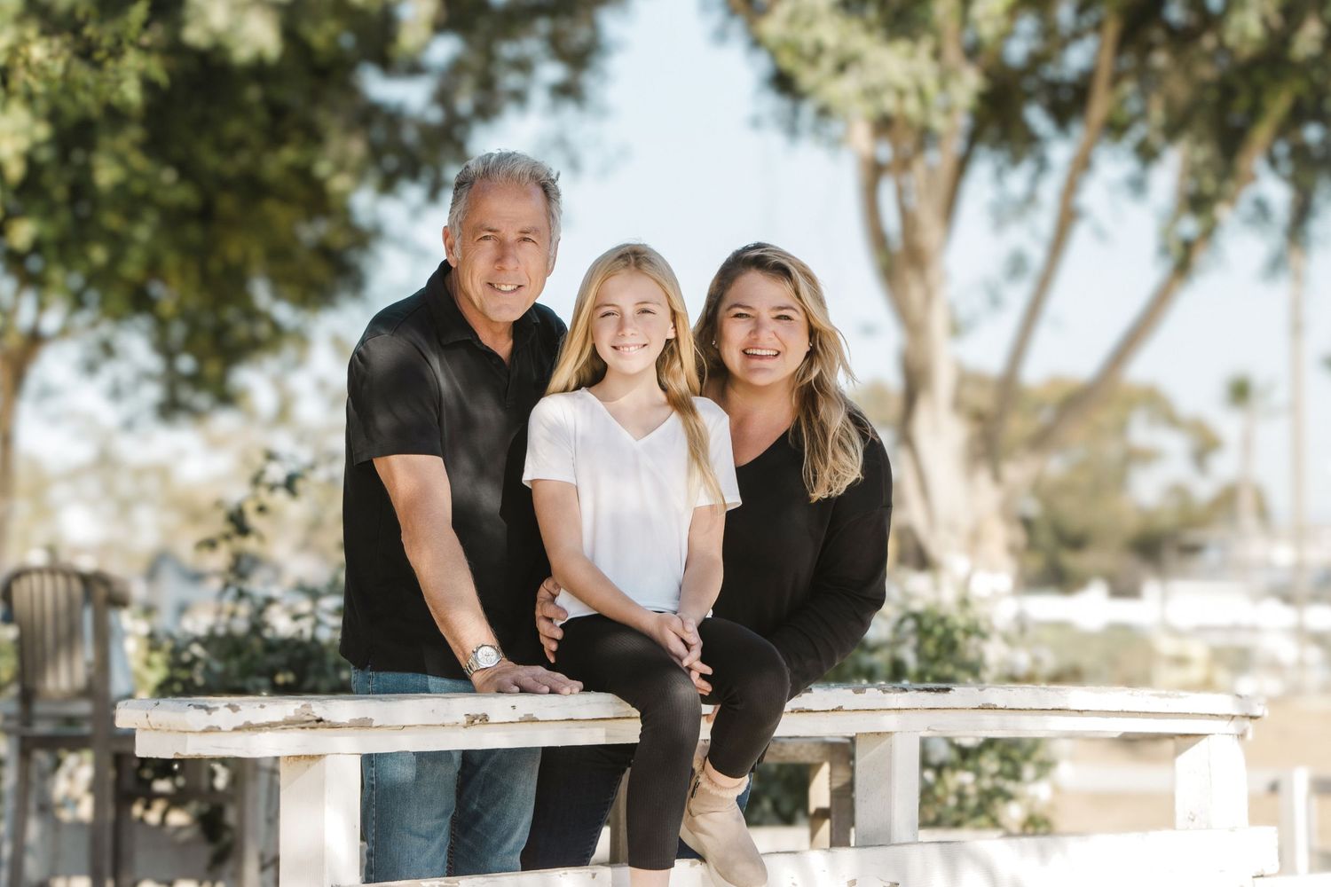 Family of three smiling outdoors on a white bench surrounded by trees in a sunny park setting.