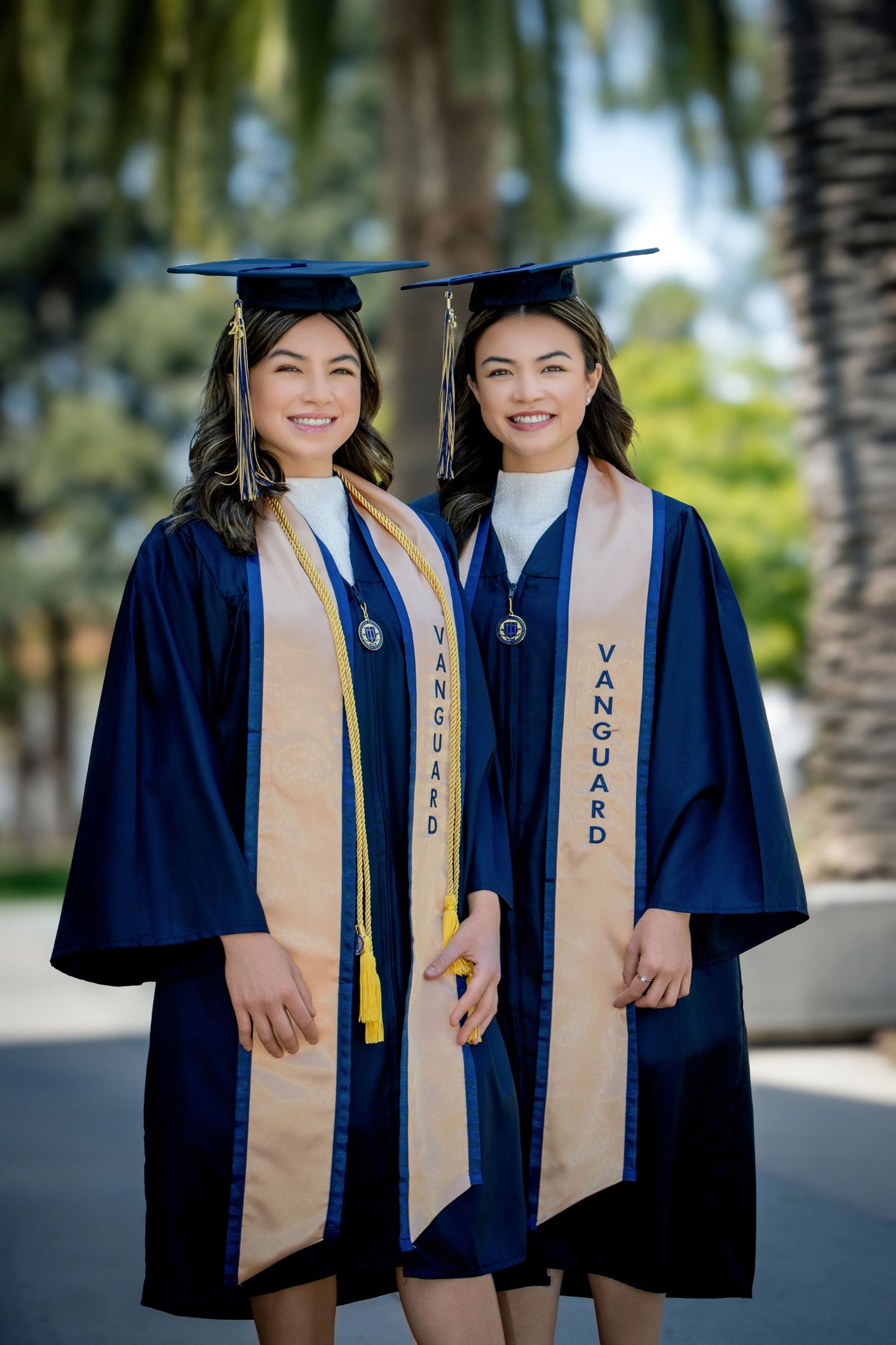 Two smiling women in navy Vanguard graduation gowns and caps pose outdoors with palm trees in the background.