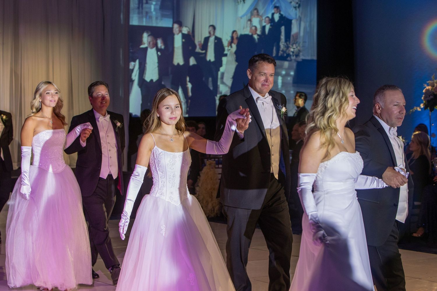 Elegantly dressed couples dance at a formal gala, wearing ball gowns and tuxedos under a projected backdrop.