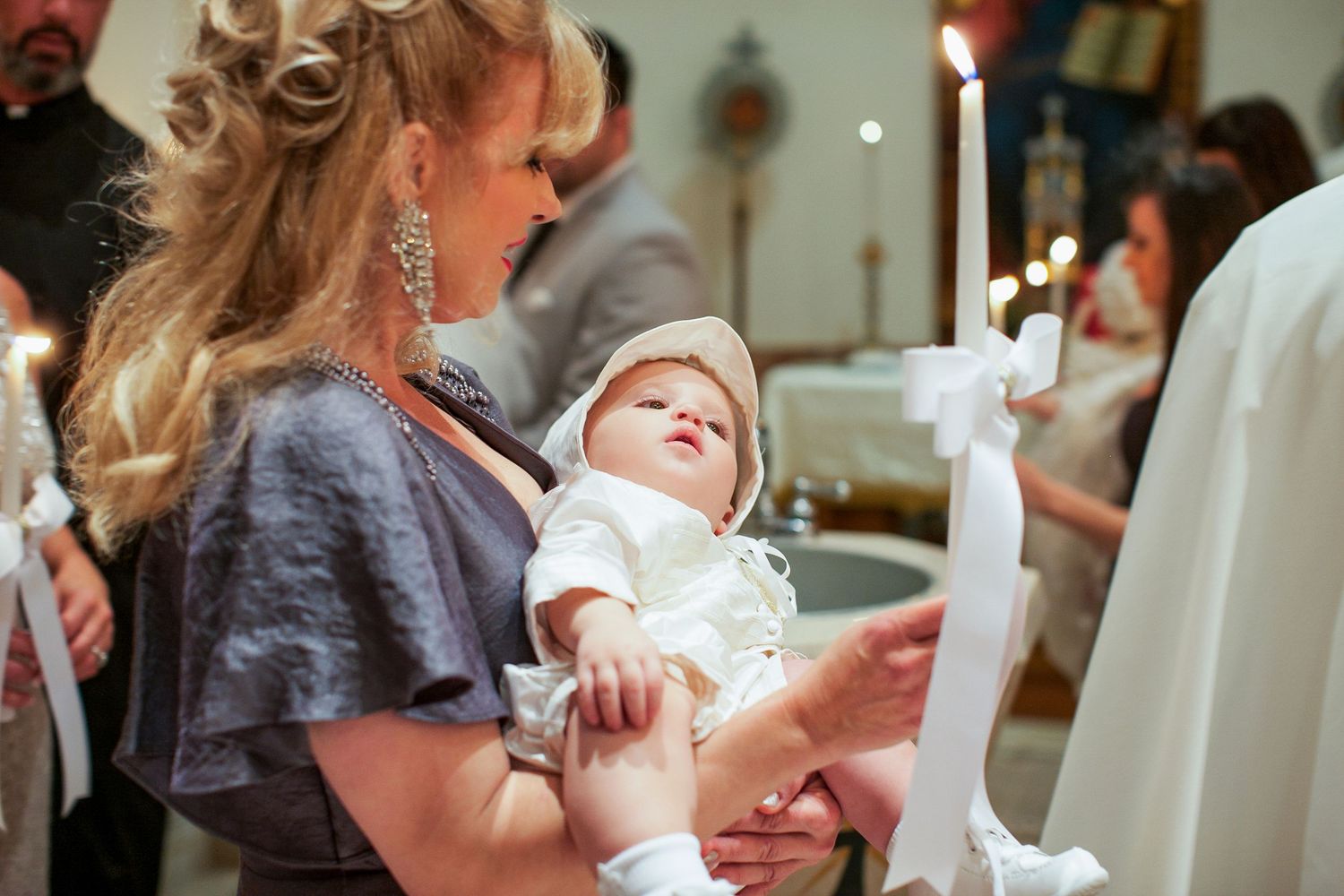 Woman in elegant gray dress holds infant in white christening gown near candles during baptism ceremony.