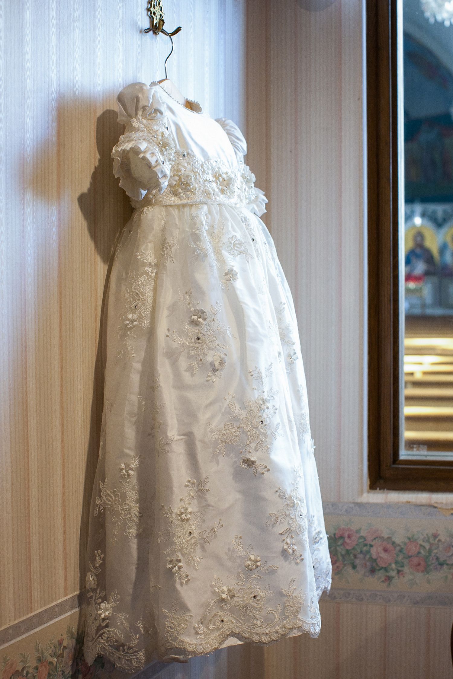 Elegant white lace christening gown with floral embroidery displayed on a mannequin near a window.