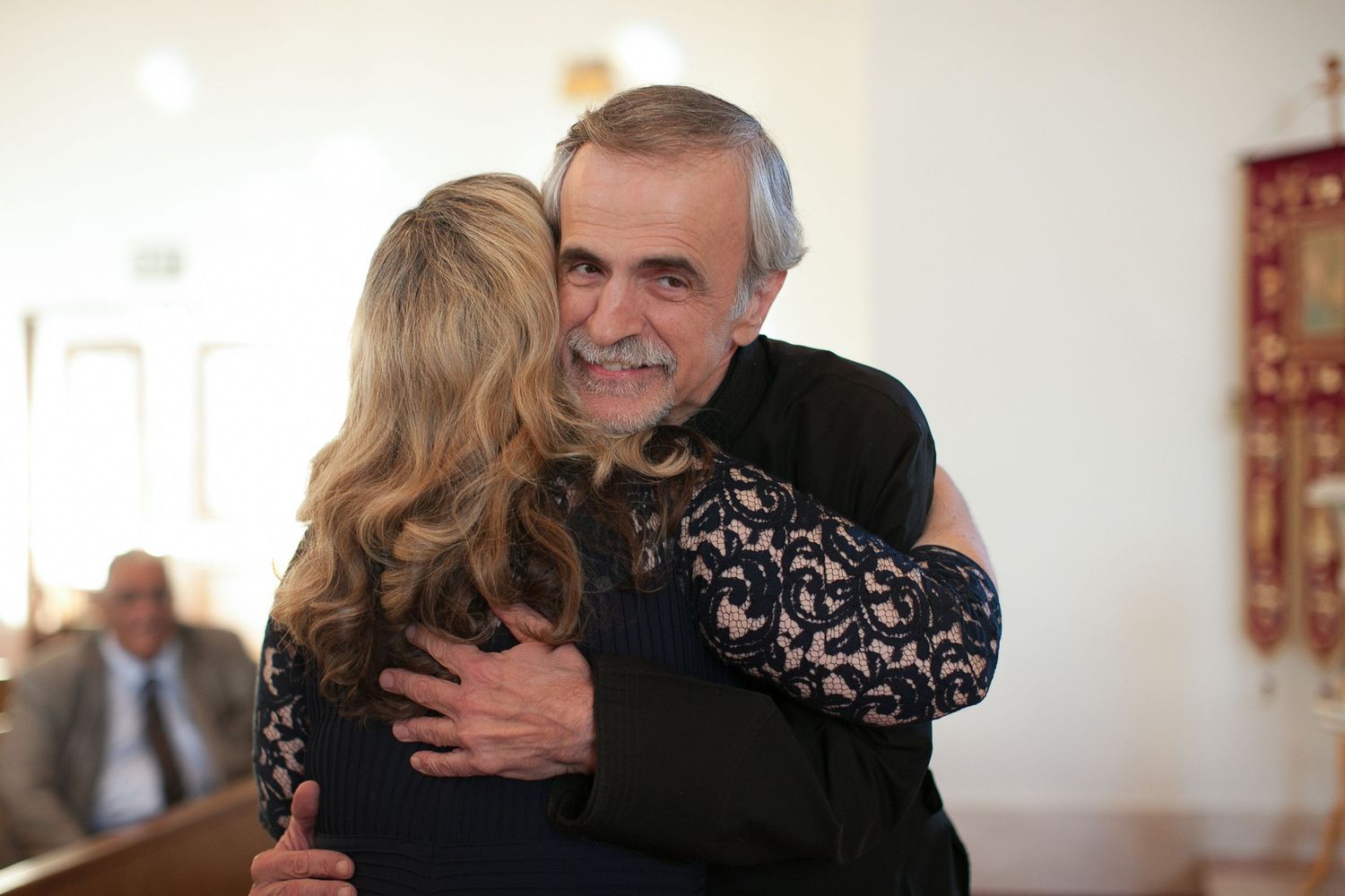 Two people sharing a warm hug in a bright room, with a smiling man in a dark lace shirt embracing a blonde woman.