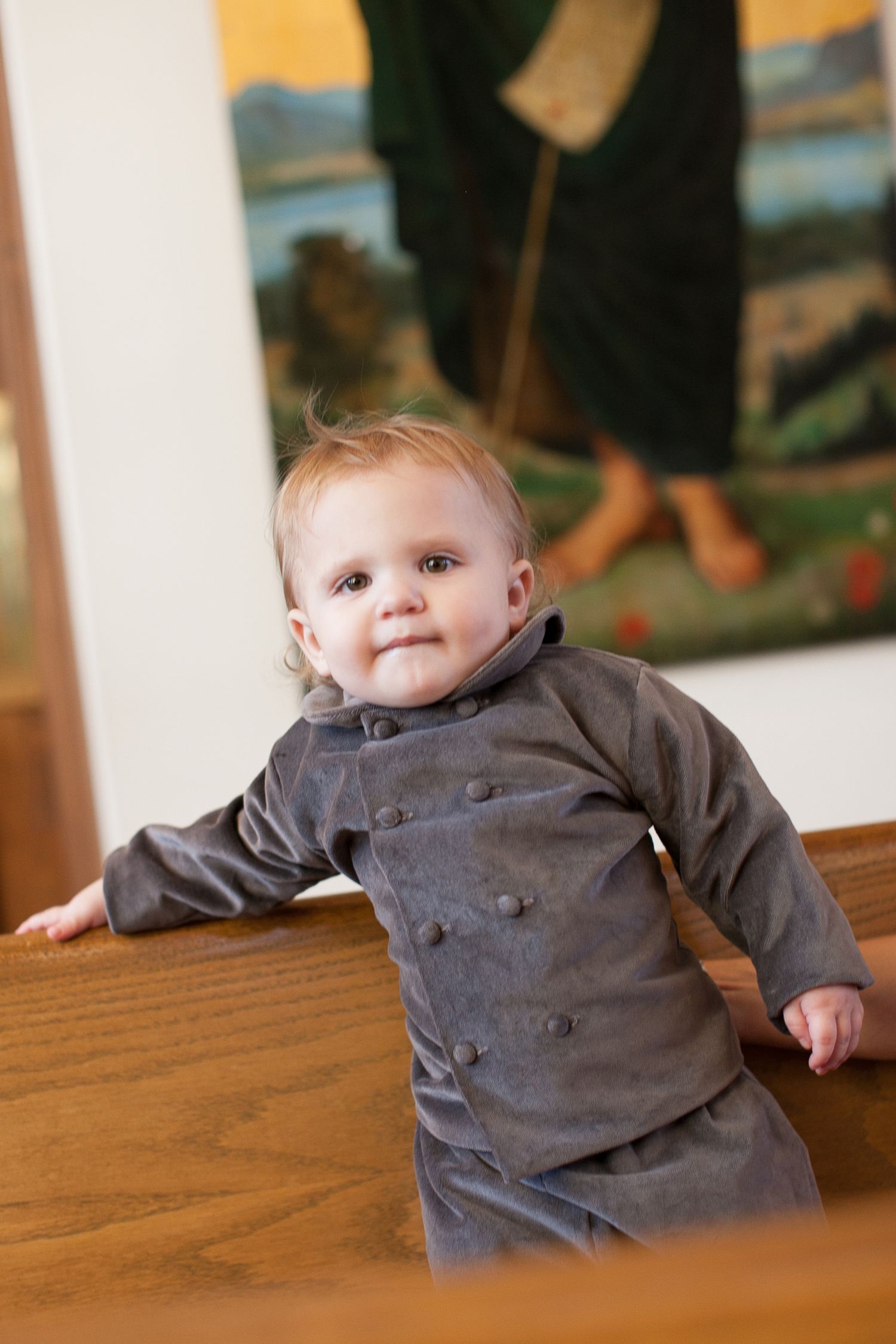 Adorable baby in gray velvet double-breasted outfit standing at wooden bench with painting in background.