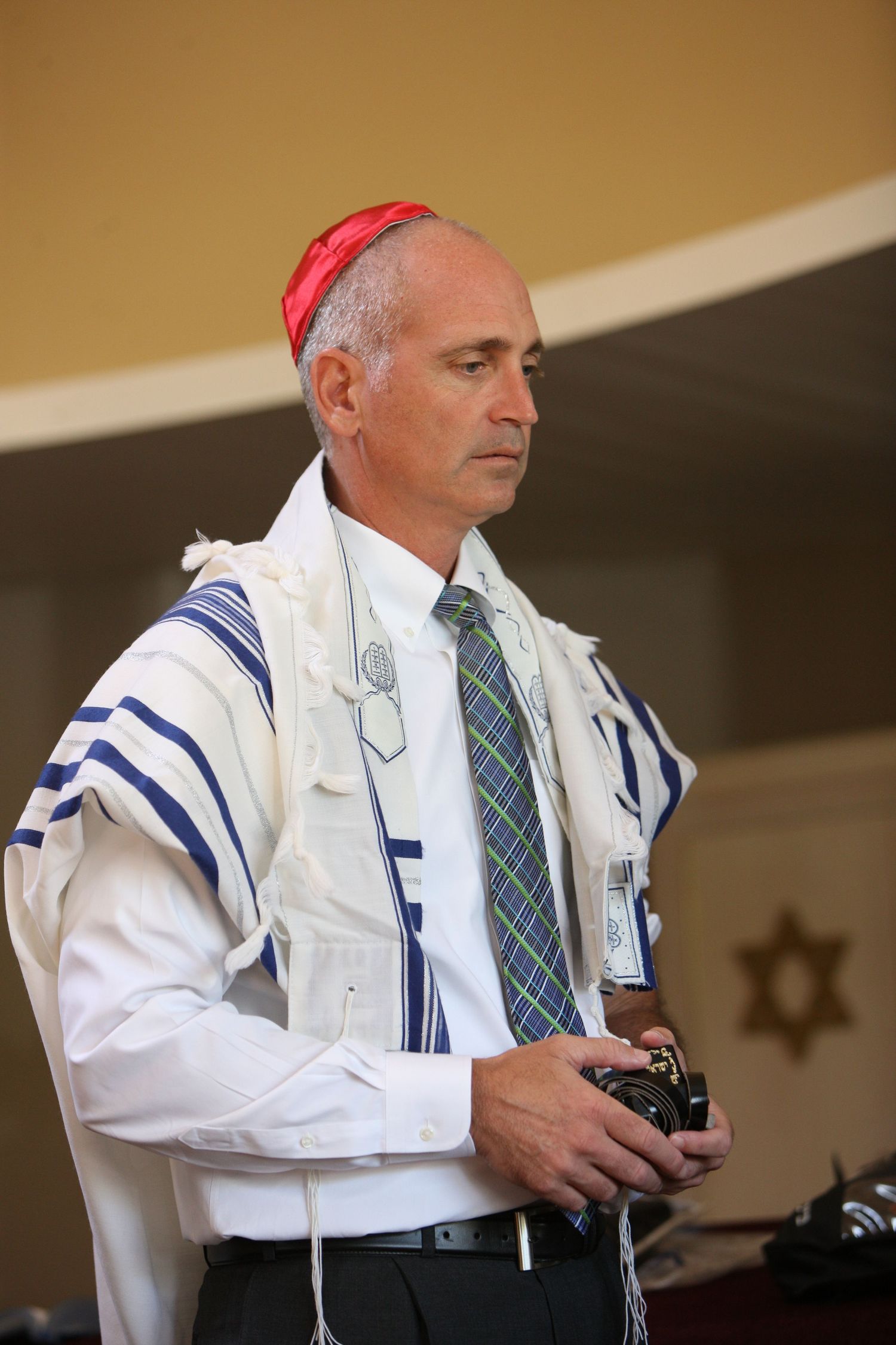 Jewish man wearing tallit and kippah holds camera during religious ceremony in synagogue.