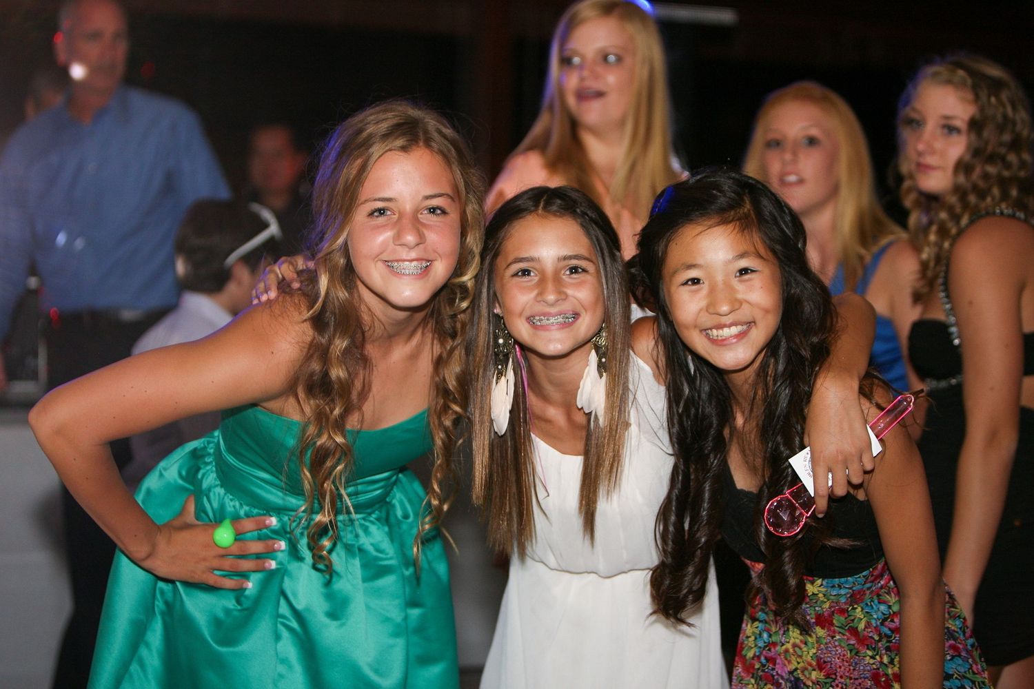 Three smiling teenage girls posing together at a party, wearing colorful dresses in green, white, and floral print.