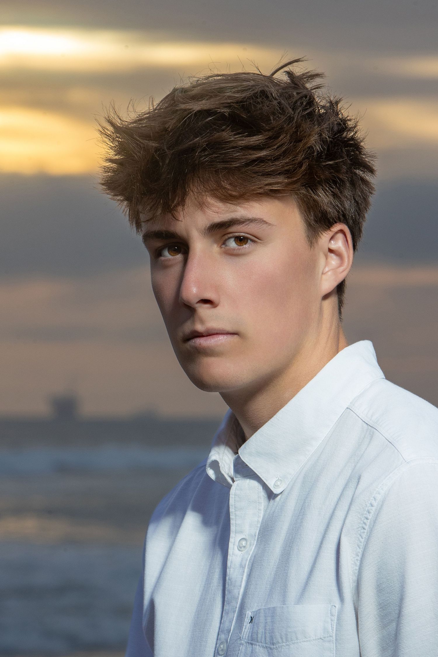 Young man with windswept brown hair in white shirt poses by ocean at sunset.