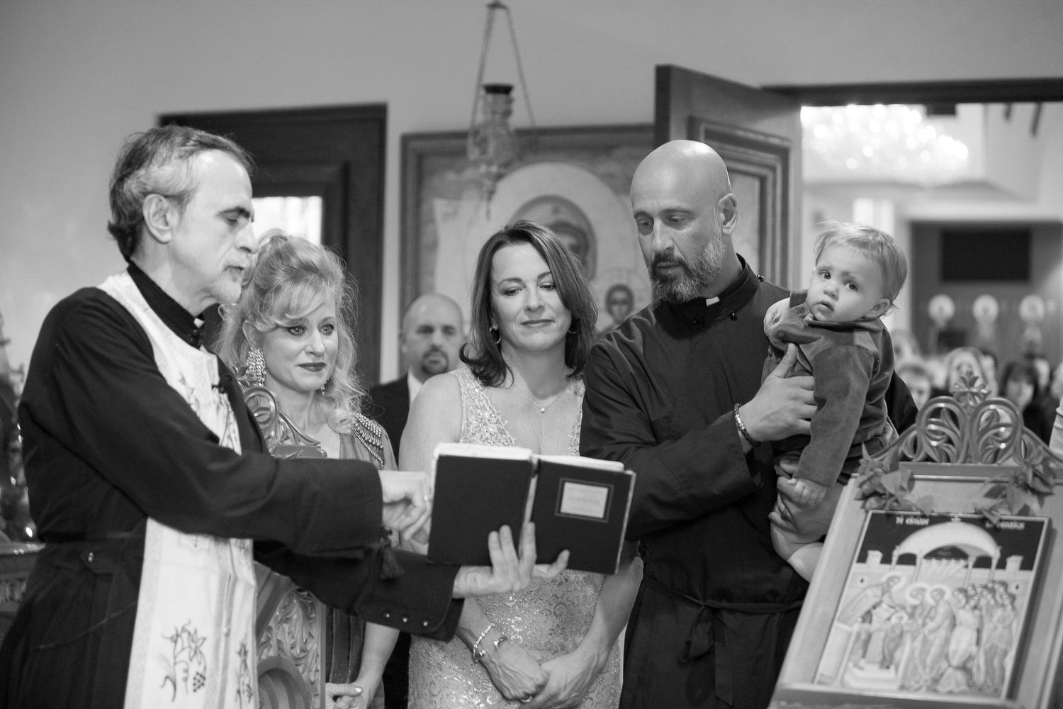 Black and white photo of a priest reading from a book during a baptism ceremony with family members present.