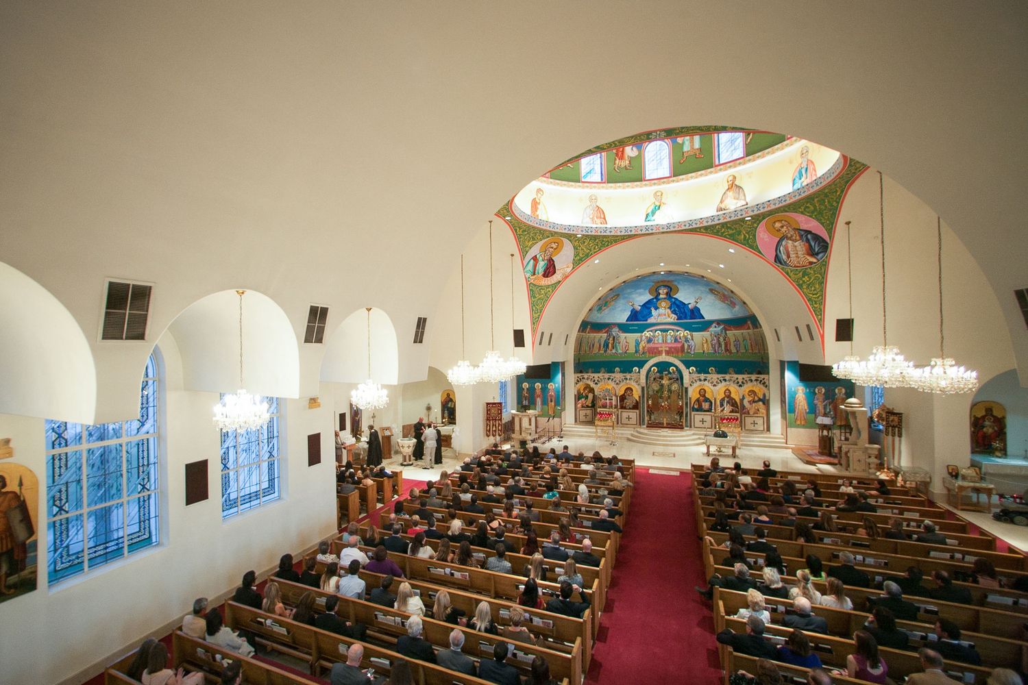Grand Orthodox church interior with ornate Byzantine murals, packed congregation, red carpet aisle, and domed ceiling artwork.