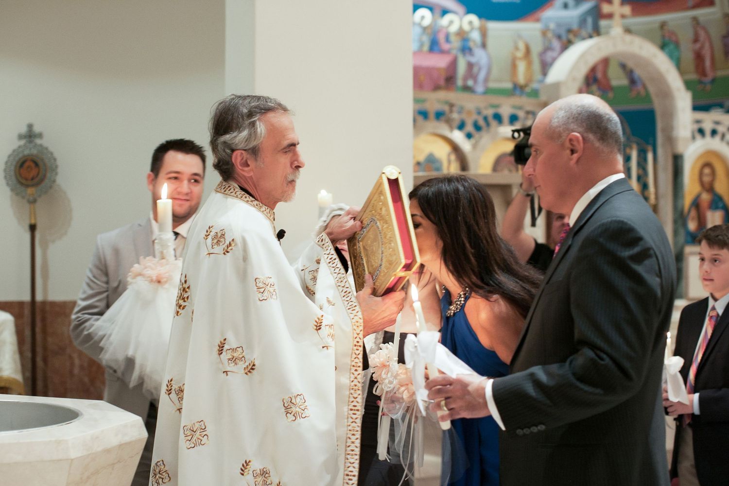 Orthodox priest in white vestments performs a religious ceremony with candles in a decorated church.