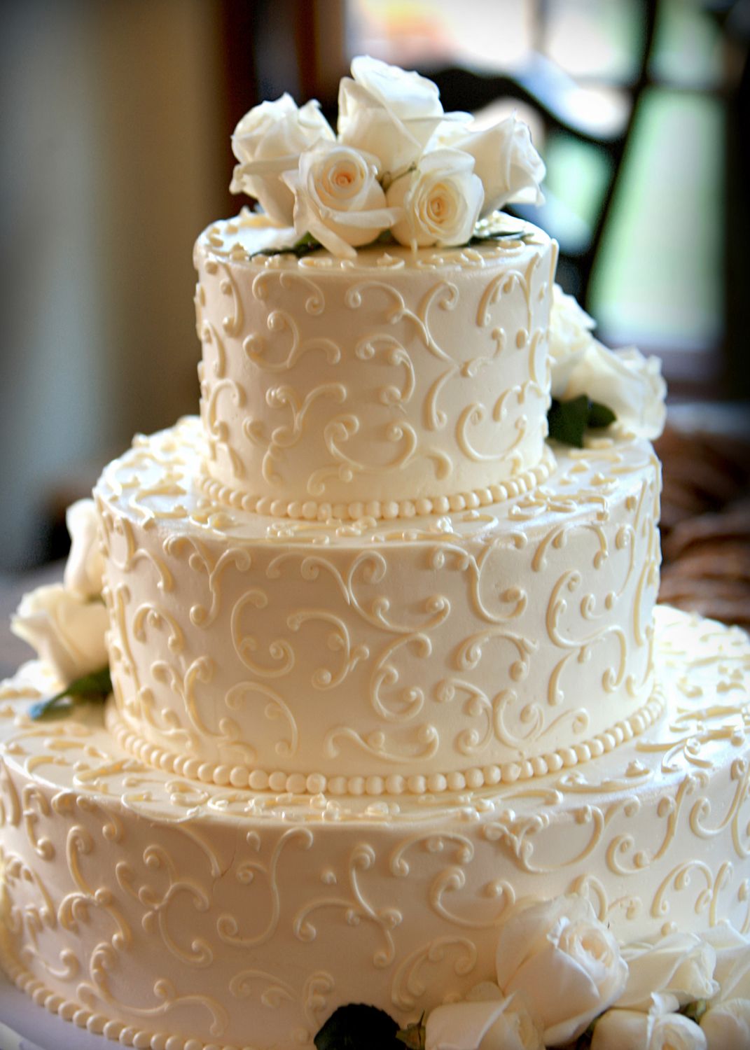 Elegant three-tier white wedding cake with intricate scrollwork frosting and fresh white roses on top.