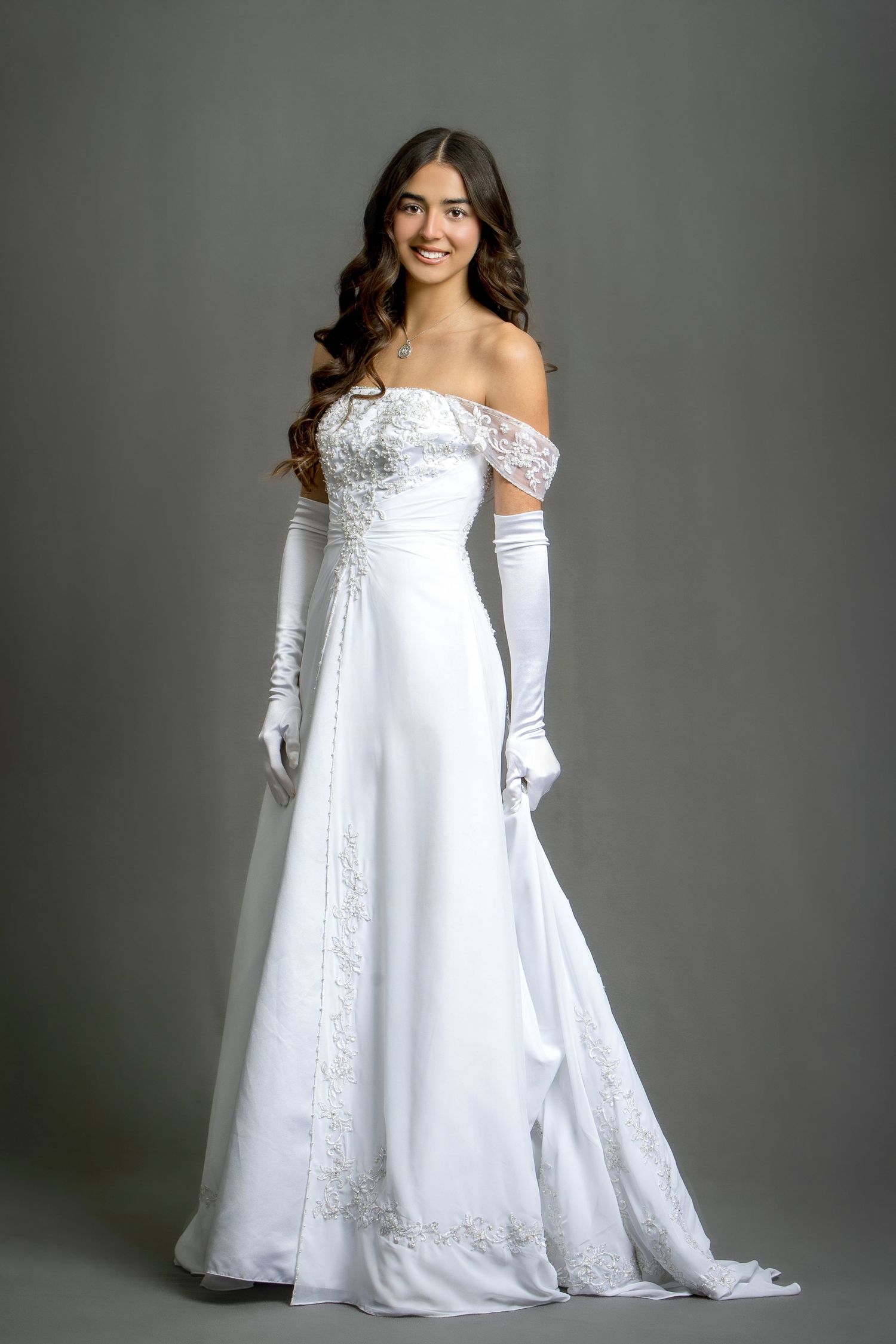 Young woman in elegant white off-shoulder wedding gown with lace details and long white gloves, smiling gracefully.