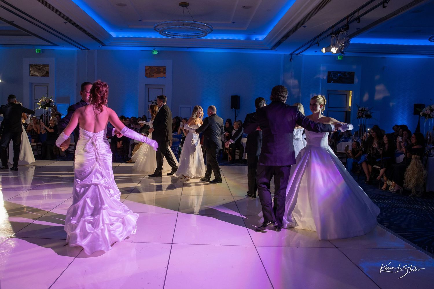 Elegant wedding reception with couples dancing on a white dance floor under blue and purple lights in a grand ballroom.