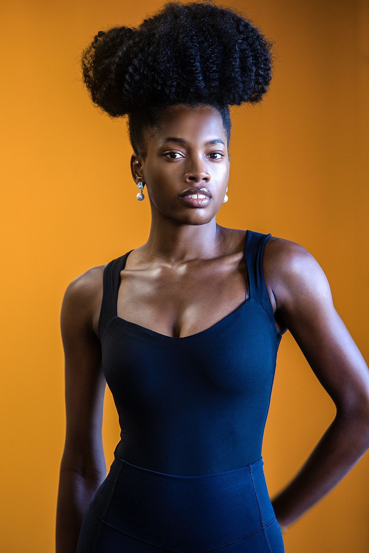 Confident Black woman with afro updo in navy bodysuit poses against vibrant orange background.