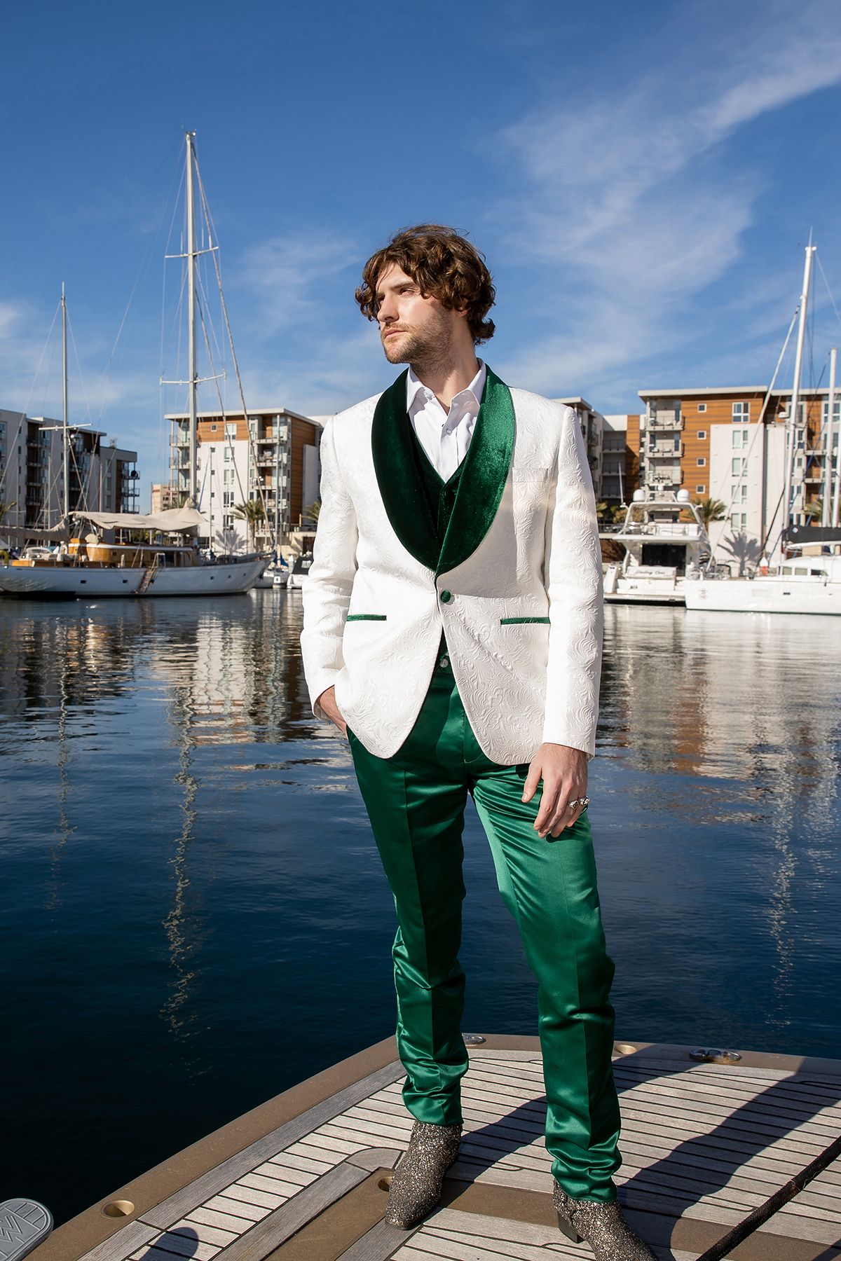 Man in white and green tuxedo suit standing on marina dock with sailboats and waterfront buildings behind him.