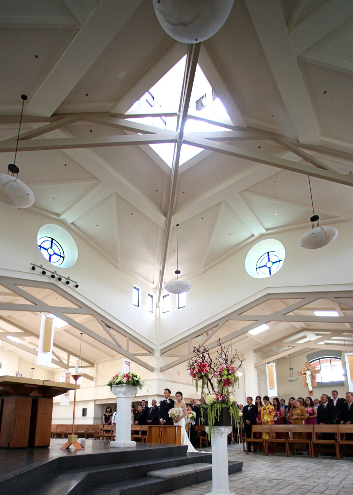 Wedding ceremony inside a modern church with soaring white vaulted ceilings, round stained glass windows, and floral arrangements.