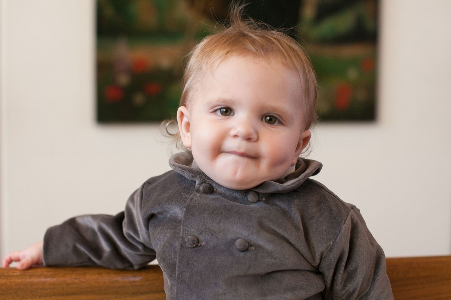 Adorable smiling baby in gray velvet outfit sitting up, with a colorful painting on the wall behind them.