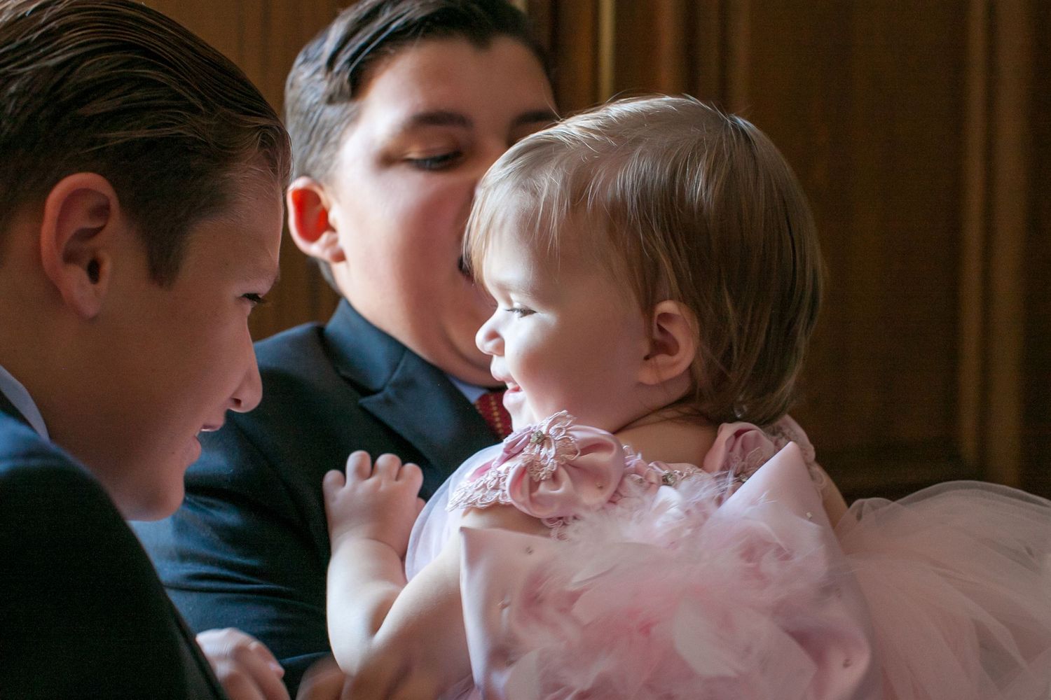 Two boys in formal suits tenderly holding a baby girl in a pink dress at a special occasion.