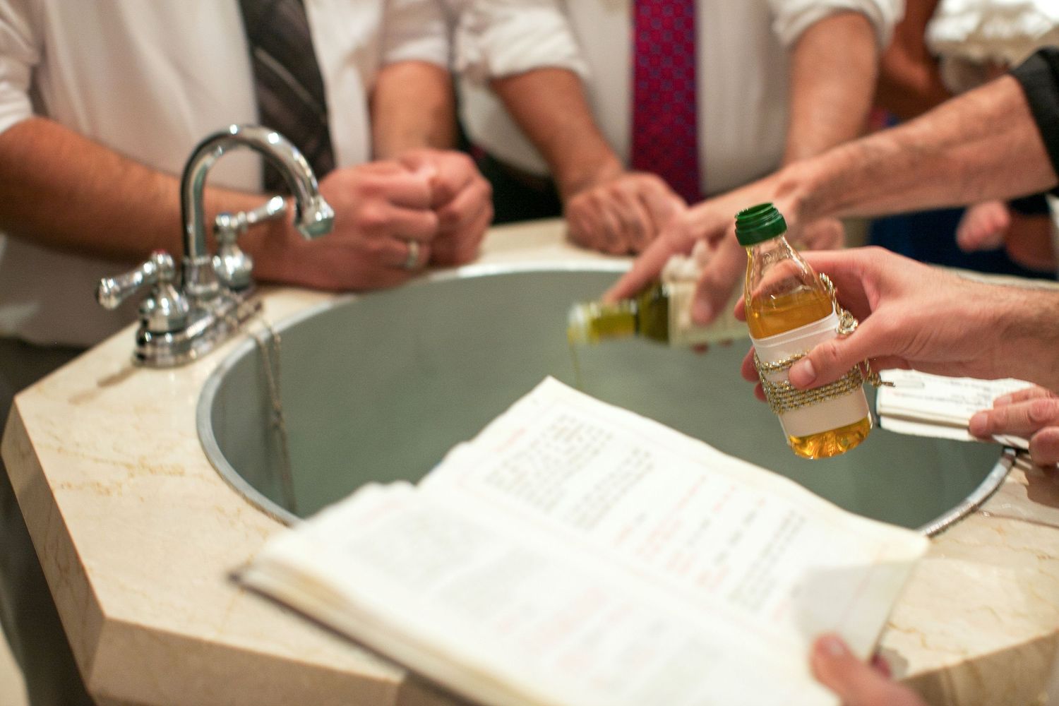 Men gathered around a sink holding a small liquor bottle and reading from a book during a formal event.