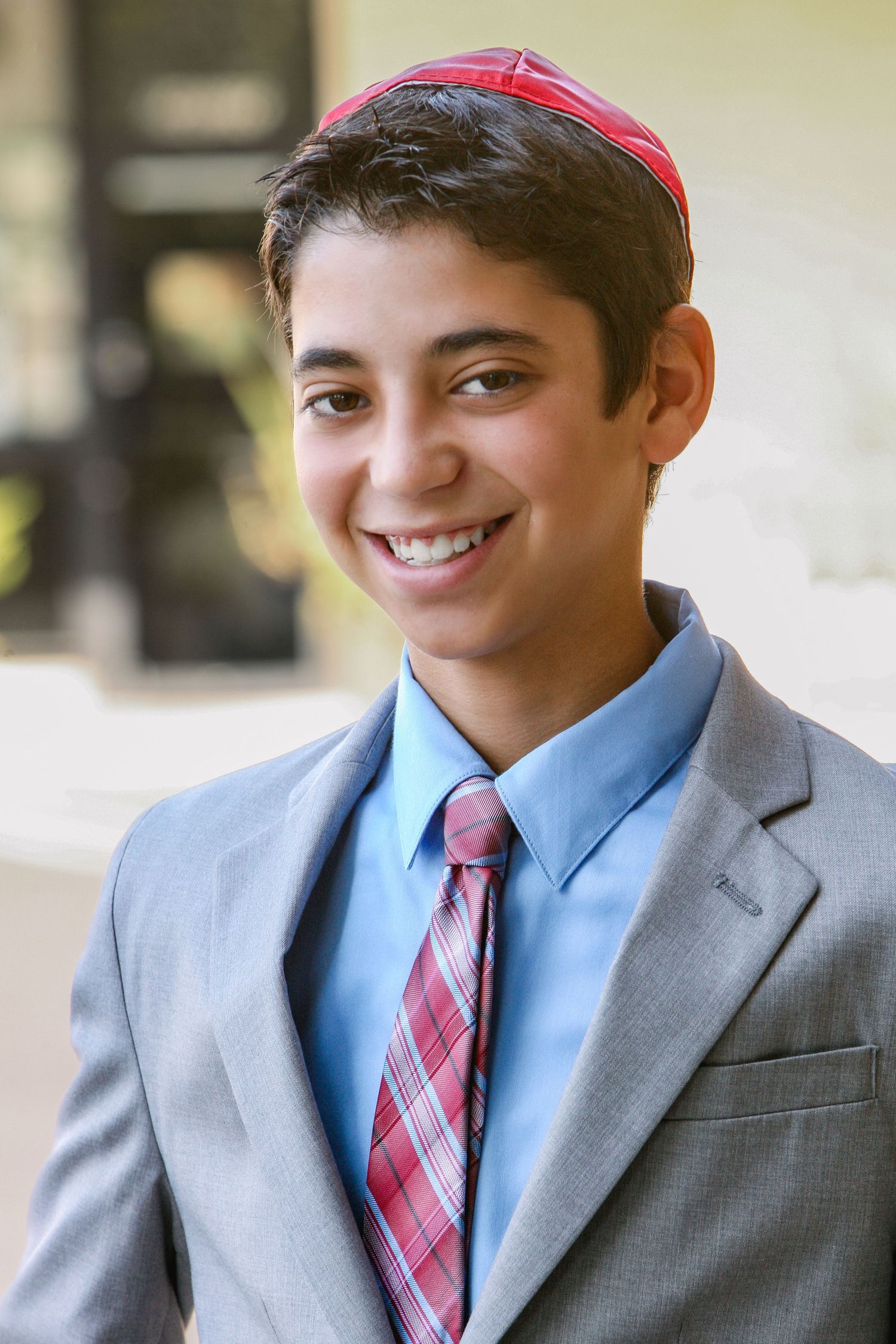 Smiling teenage boy in gray suit, plaid tie, and red kippah posing outdoors for Bar Mitzvah portrait.