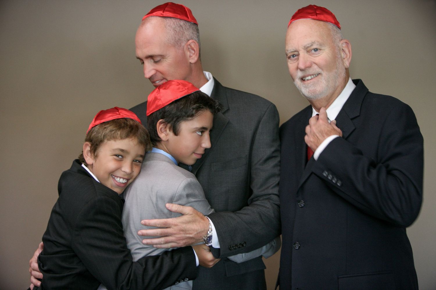 Four people wearing red kippot pose together smiling, two adults and two boys hugging at a Jewish celebration.