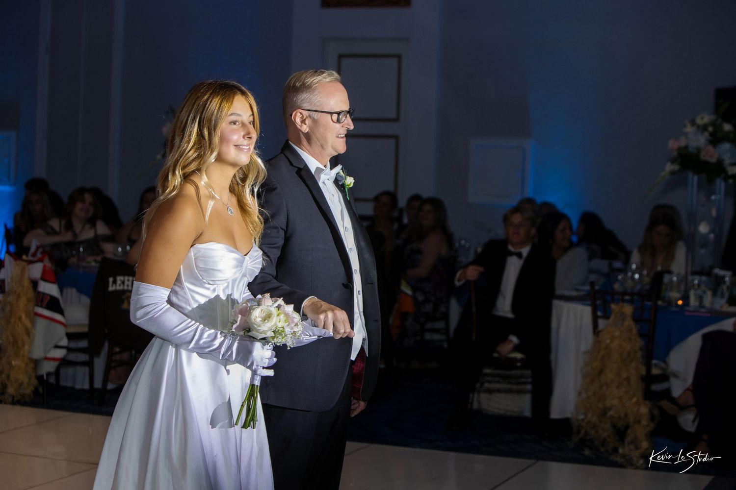 Bride in white strapless gown holding bouquet walks with escort in tuxedo at elegant wedding reception.
