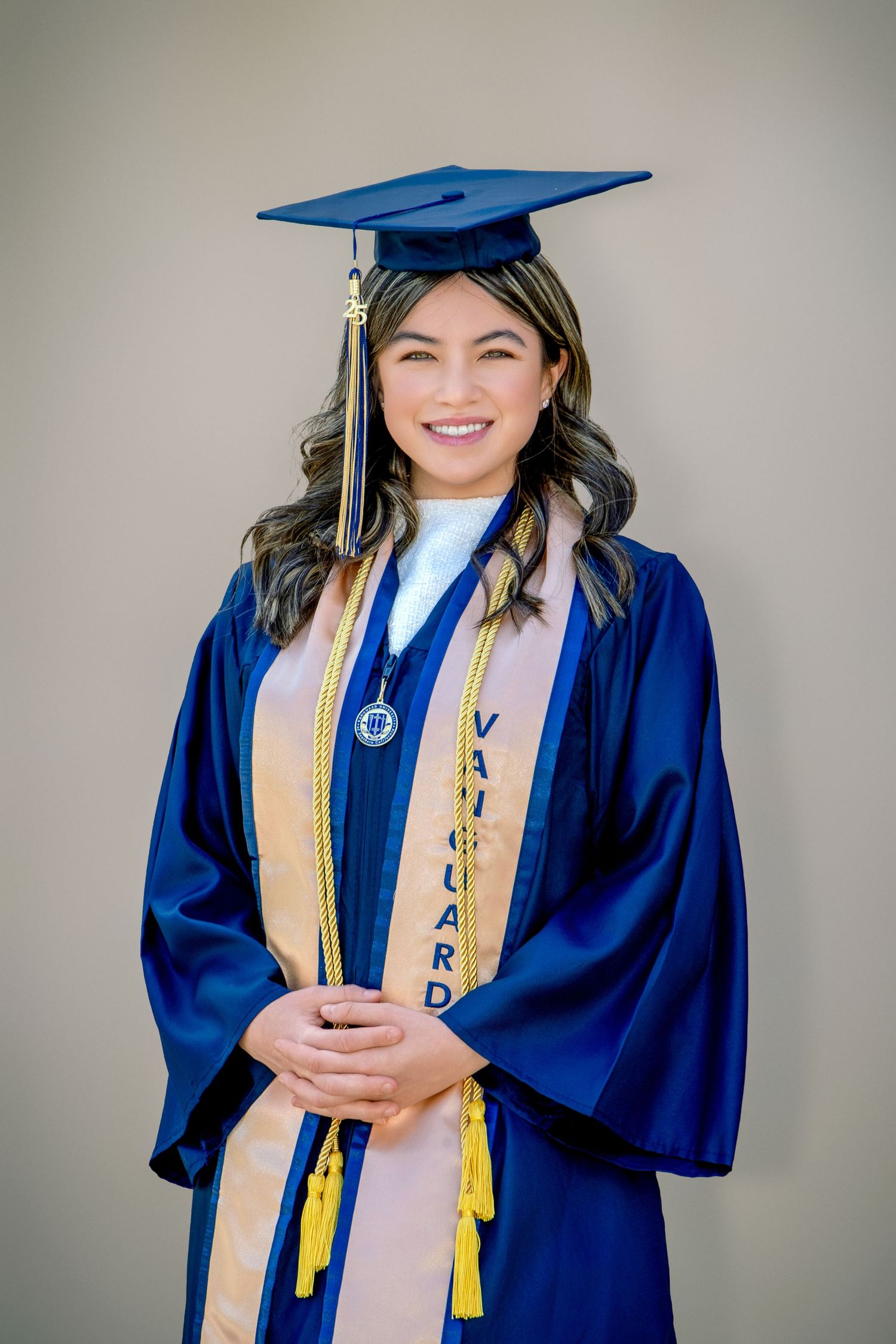 Smiling graduate in blue cap and gown with Vanguard University stole and gold honor cords poses for portrait.