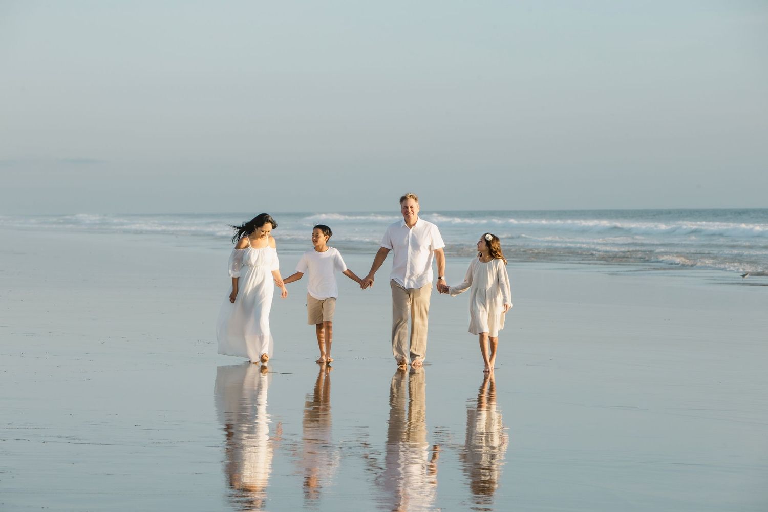 Family of four in white outfits walking hand-in-hand on a sunny beach with calm ocean waves.