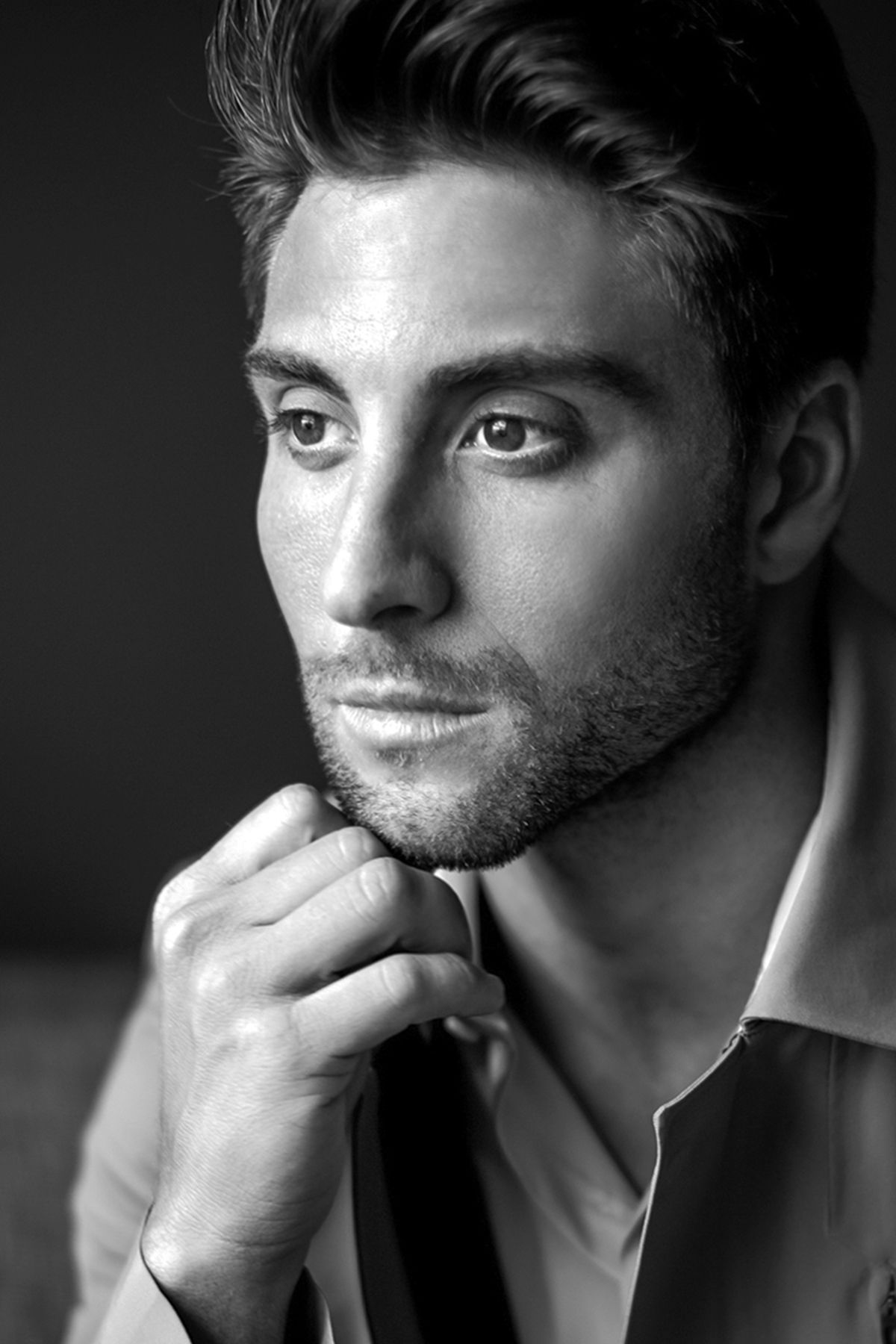 Black and white portrait of a handsome young man with stubble, adjusting his tie while gazing thoughtfully.