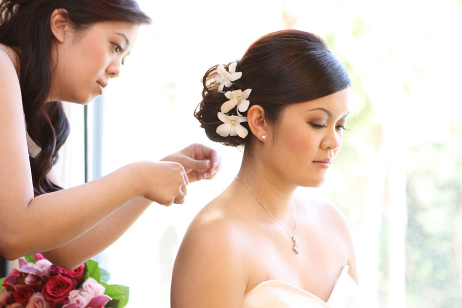 Bridesmaid helping bride with necklace while bride wears white dress and floral hair accessories.