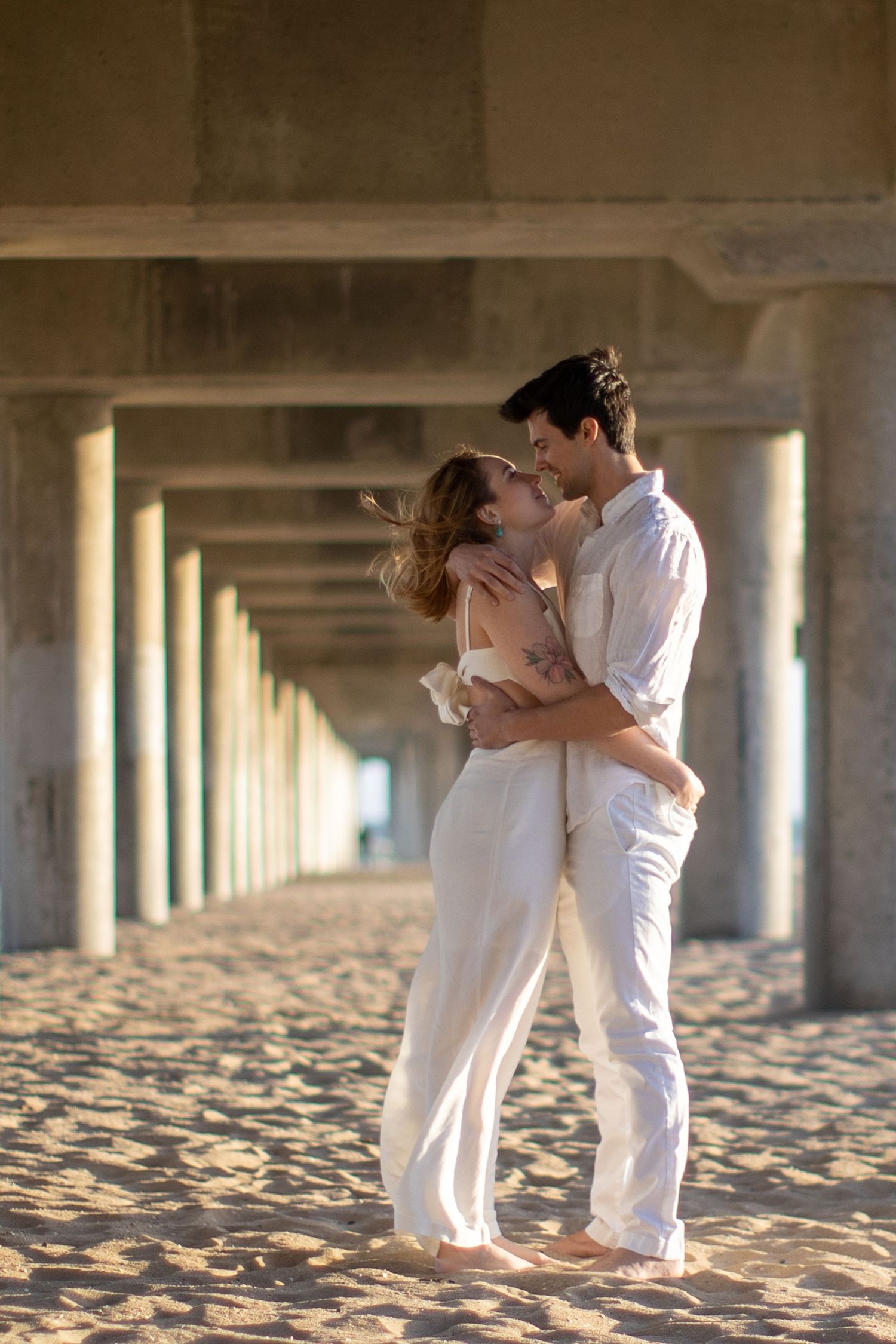 Loving couple in white outfits embrace and kiss barefoot on sandy beach under pier columns.
