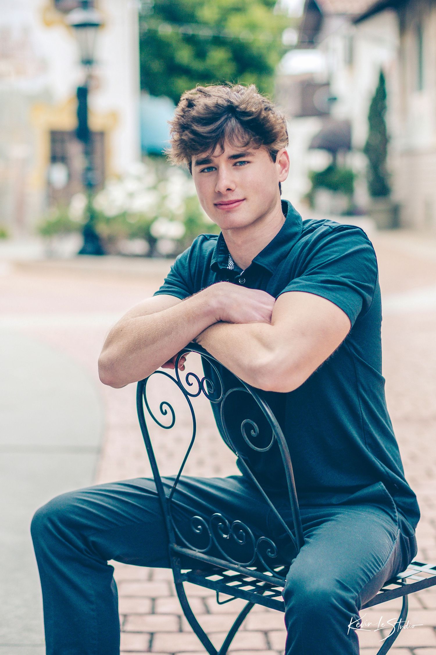 Handsome young man in teal polo shirt sitting on ornate iron bench on a brick walkway.