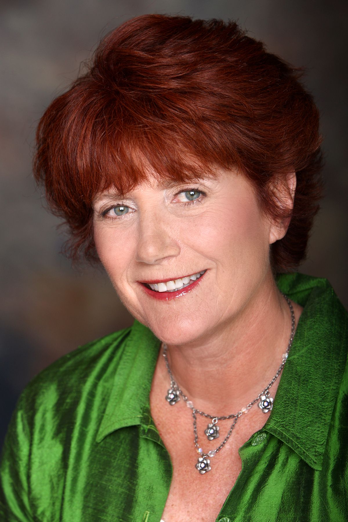 Professional headshot of a smiling middle-aged woman with short red hair wearing a green blouse and silver necklace.