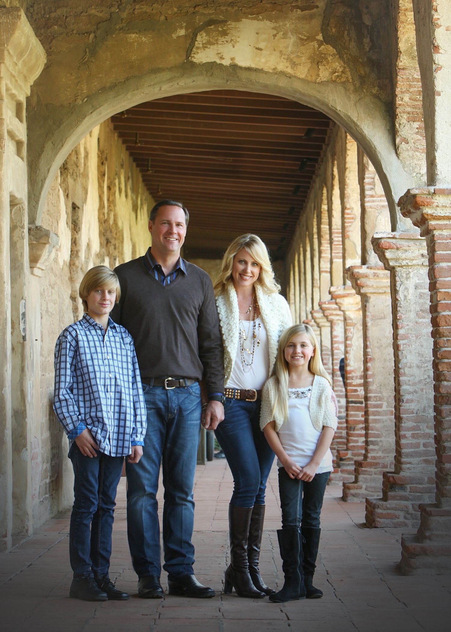 Family of four posing under historic brick arched corridor, parents with two children smiling in casual western attire.
