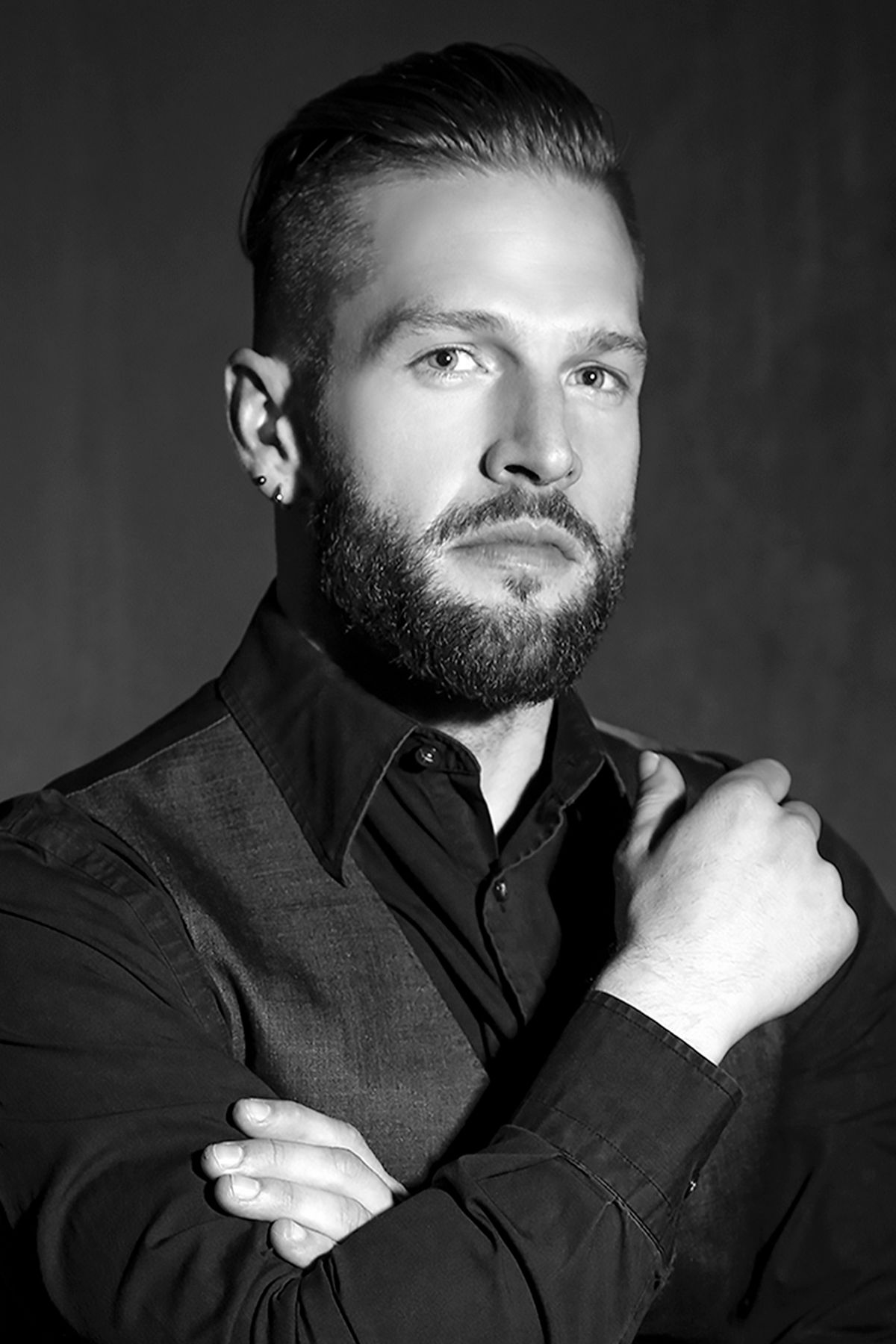 Black and white studio portrait of a bearded man in dark shirt with arms crossed, looking confidently at camera.