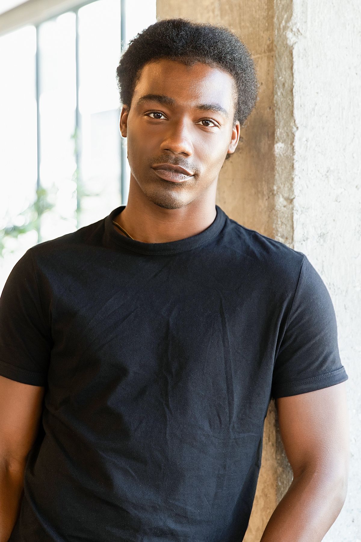Young Black man with short hair wearing a plain black t-shirt, standing against a concrete wall.