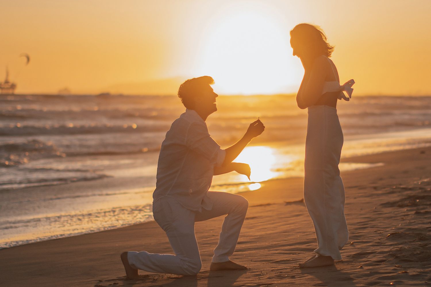 Romantic beach proposal at sunset with man on one knee presenting ring to his partner.