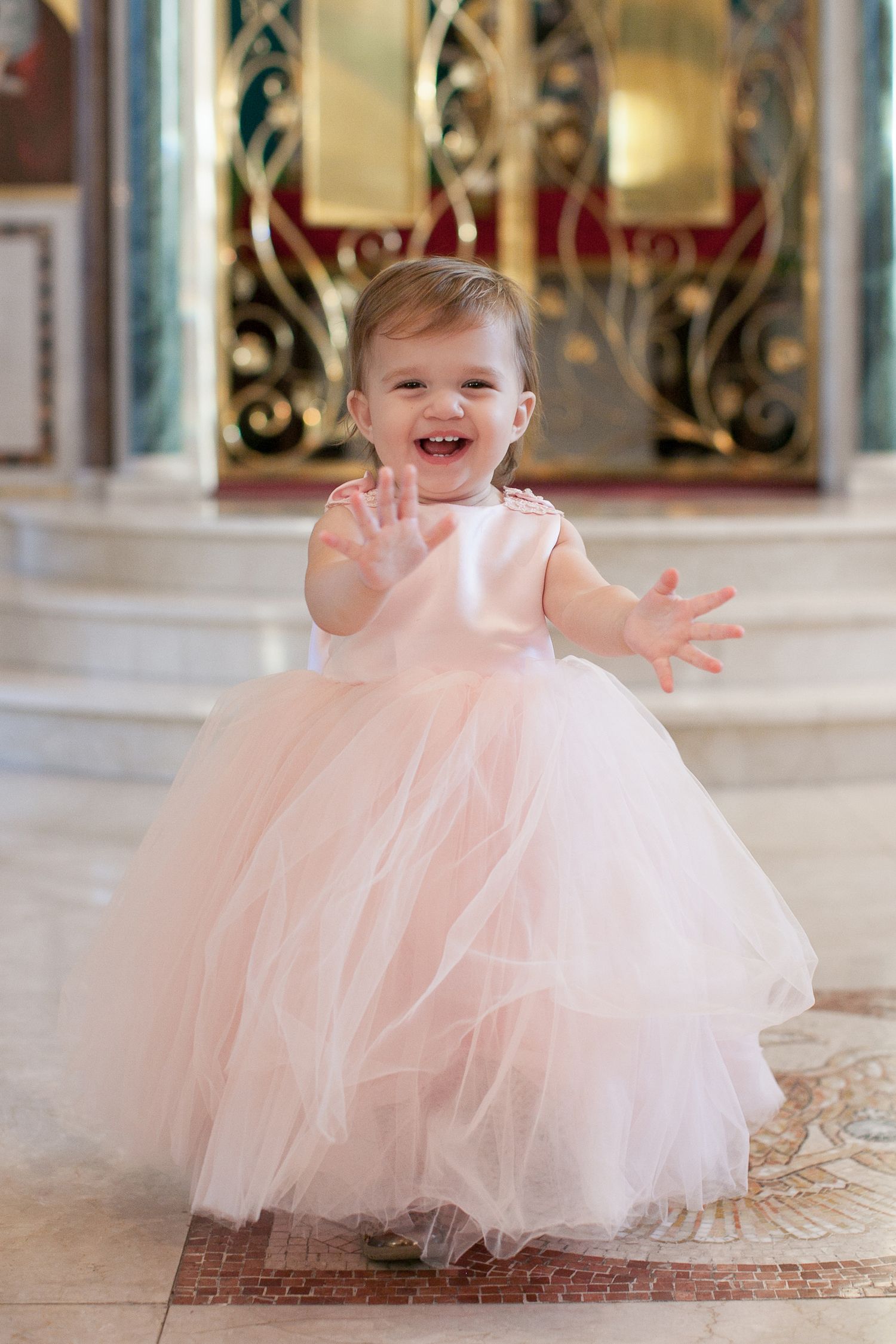 Adorable baby girl in a pink tulle ball gown laughing and reaching out her hands in an elegant venue.