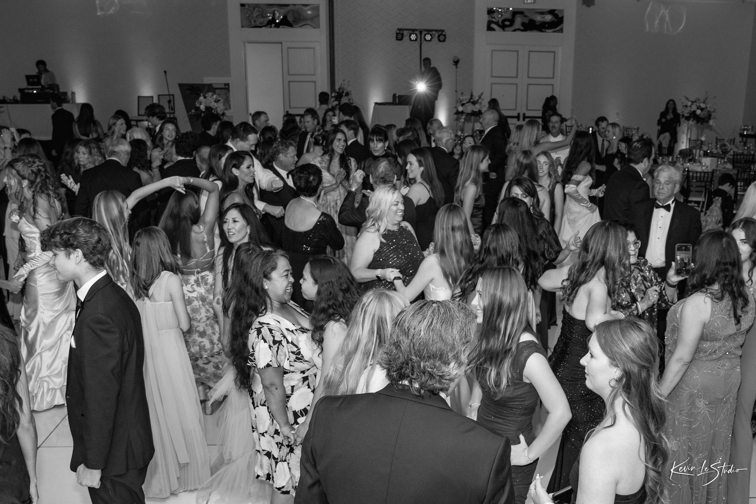 Black and white photo of elegantly dressed guests dancing at a formal wedding reception in a grand ballroom.