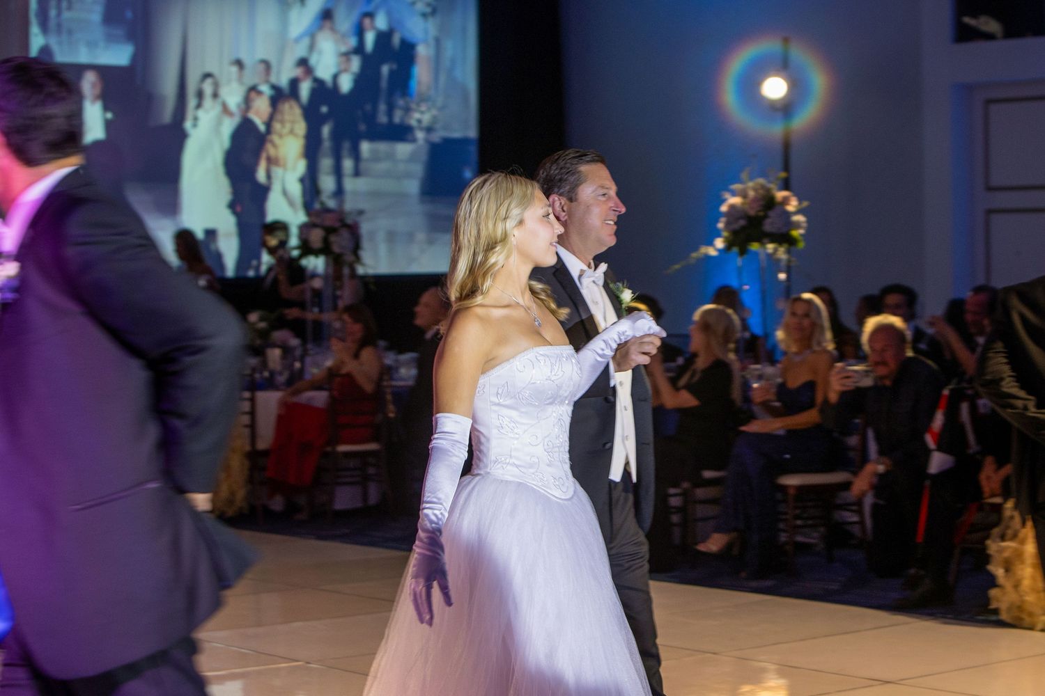 Elegant couple dancing at a formal gala event, woman in white ballgown with gloves, man in tuxedo, ballroom setting.