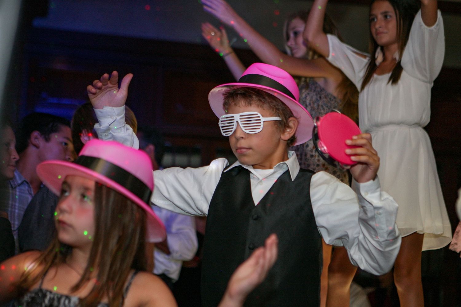 Children dancing at a party wearing pink fedora hats and novelty shutter shades glasses at a festive celebration.