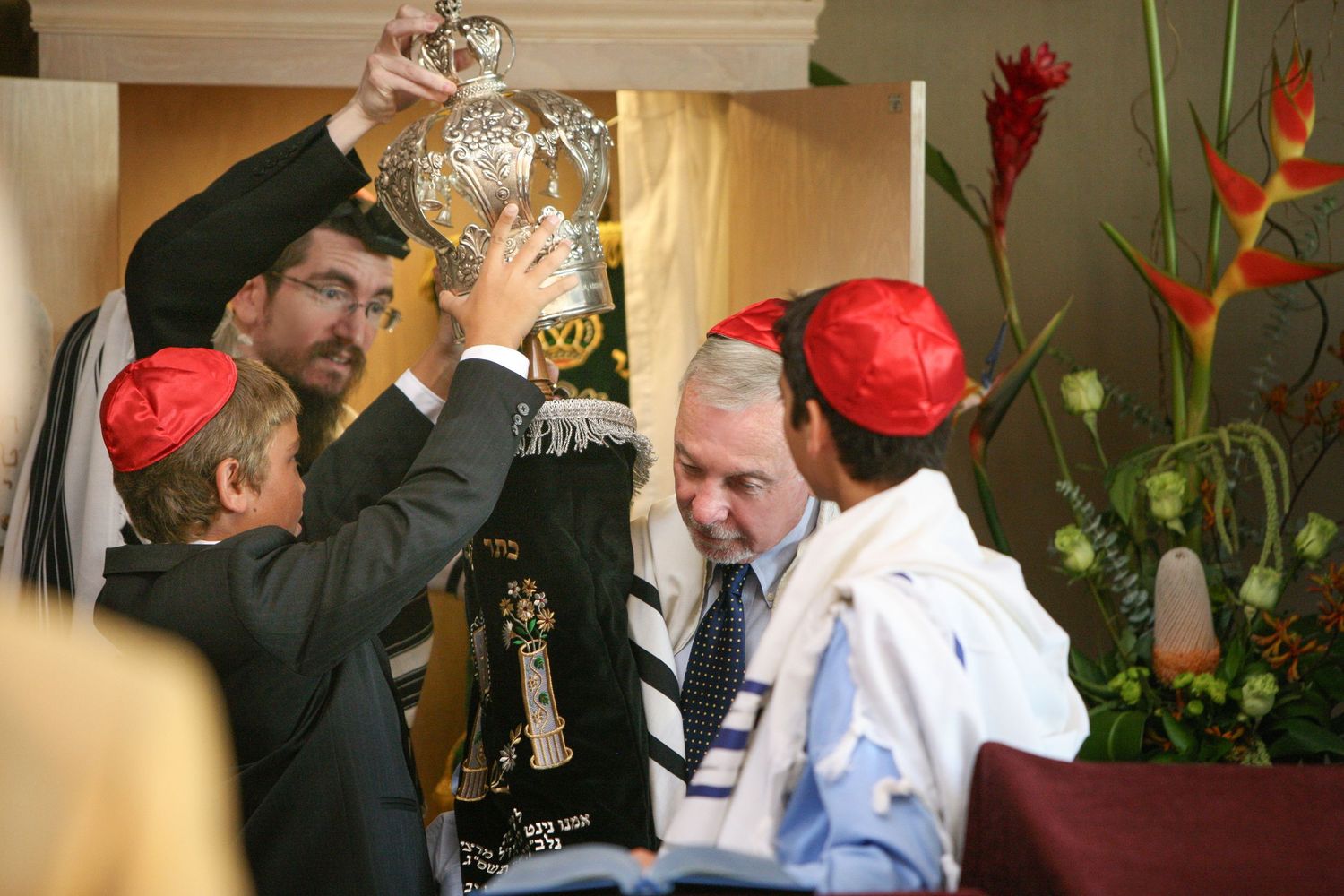 Men in red kippot and tallitot participate in a Jewish religious ceremony, lifting a Torah scroll during synagogue services.